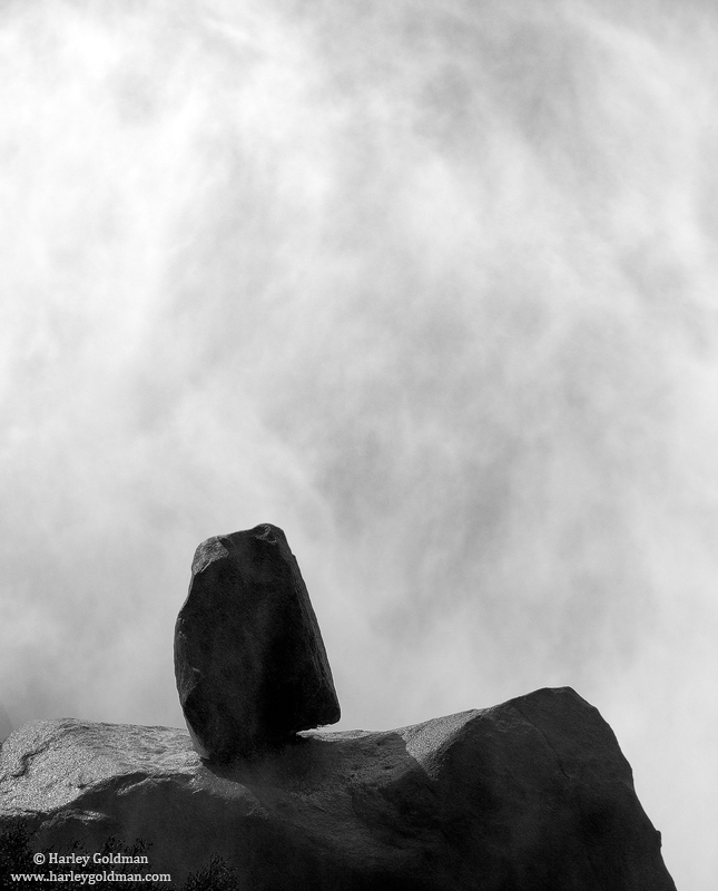 A granite boulder is balance on a bigger boulder with the spray of Nevada Falls in the background